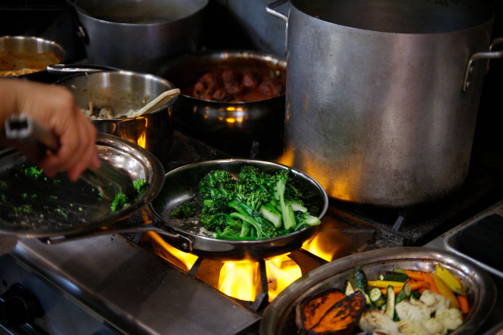 Broccolini searing in a pan over the flame, beside a tray of roasted vegetables in Solera's kitchen
